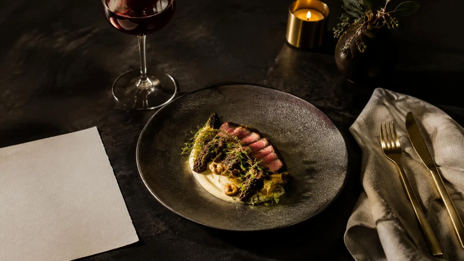 Plato de carne con colmenillas y puré sobre plato de piedra oscura, junto a una copa de vino tinto y una carta en blanco